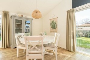 A dining area with a round table, white chairs, a cabinet with glass doors, a hanging light fixture, and a view through a window with curtains. - Holiday home in Renesse with swimming pool (Renesse)