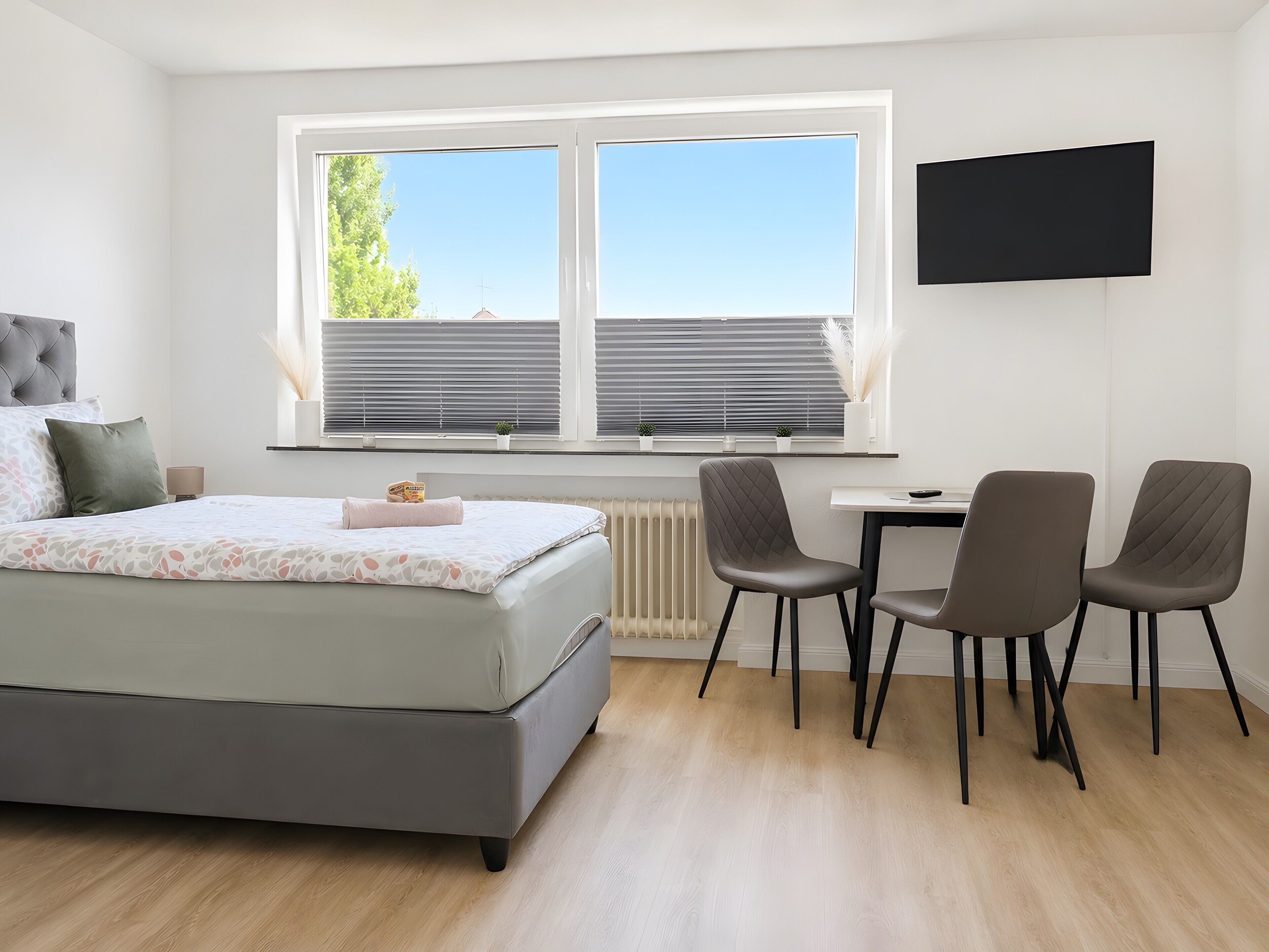 A modern bedroom with a bed, a desk, two chairs, a radiator, and a TV mounted on the wall.