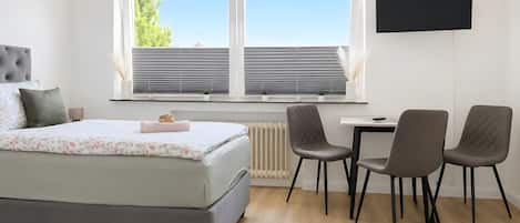Une chambre moderne comprenant un lit, un bureau, deux chaises, un radiateur et un téléviseur fixé au mur.