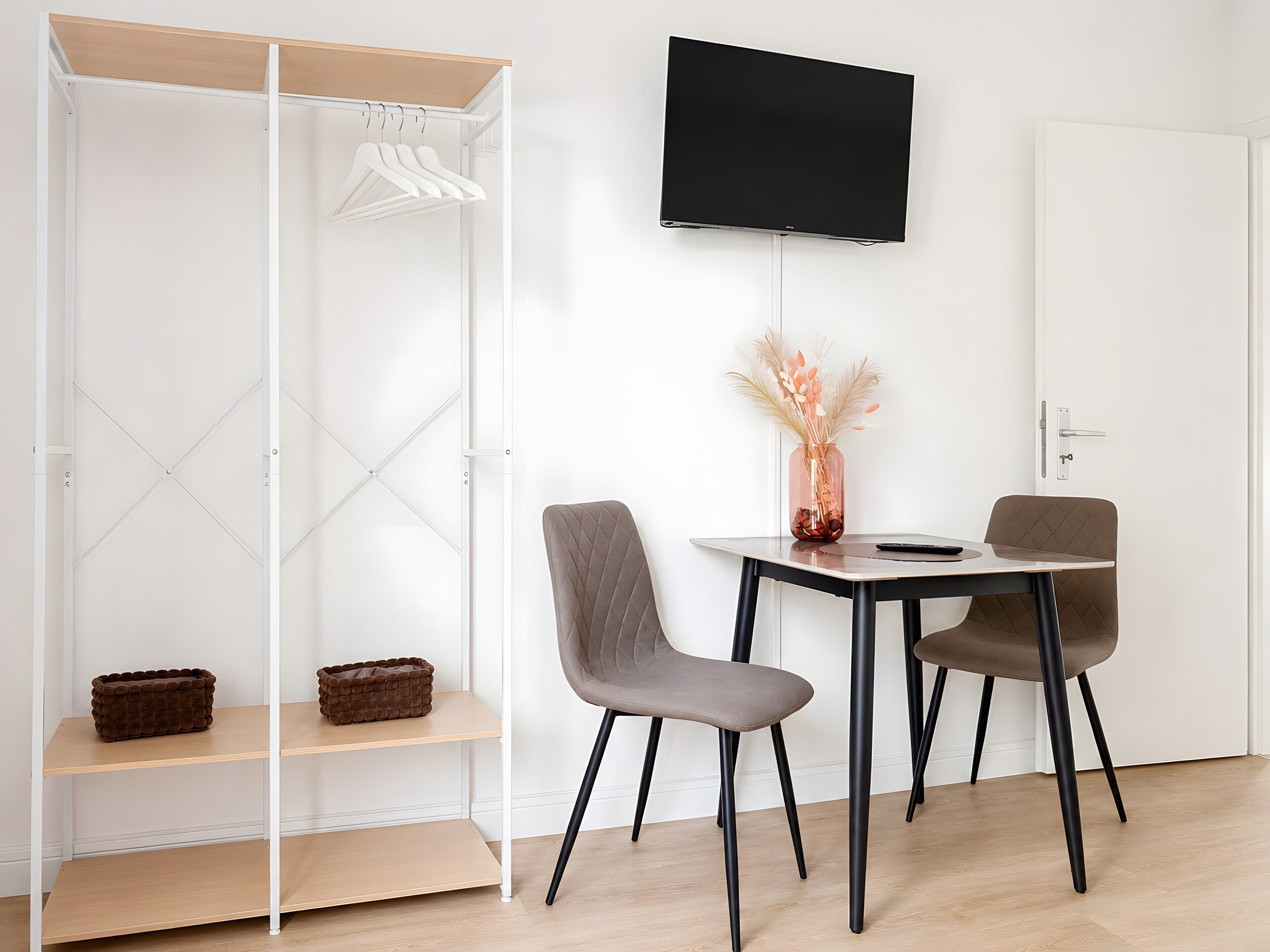 Ein modernes Zimmer mit einem weißen Kleiderschrank, einem Flachbildfernseher, einem Tisch mit zwei Stühlen und einer Vase mit Trockenpflanzen.