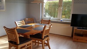 Espaço de jantar com mesa e cadeiras de madeira, televisão sobre suporte e janela com vista para áreas verdes.