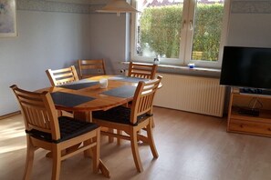 A dining area with a wooden table and chairs, a television on a stand, and a window with a view of greenery. - Holiday apartment with 2 bedrooms (Norden-Norddeich)