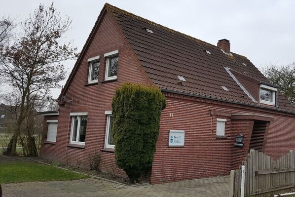 A red brick house with a sloped roof, a small garden, and a wooden fence. - Holiday apartment with 2 bedrooms (Norden-Norddeich)