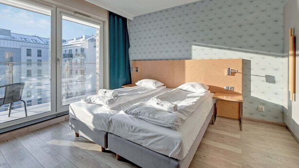 Ein modernes Hotelzimmer mit einem großen Bett, einem kleinen Nachttisch und Blick auf die Stadt durch ein großes Fenster.