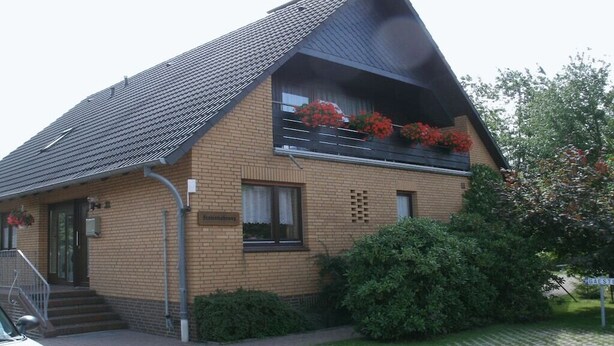 Ein zweistöckiges Gebäude mit einem geneigten Dach, einem Balkon mit roten Blumen und einer Ziegelhausfassade.
