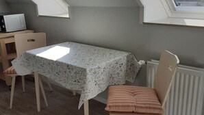 Ein Zimmer mit einem Tisch, bedeckt mit einer gemusterten Tischdecke, einem Sessel mit Kissen, einem Heizkörper und einem Fenster.
