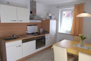 Eine kleine Küche mit Herd, Backofen und Spüle, ein Esstisch mit Stühlen und ein Fenster mit Vorhängen.
