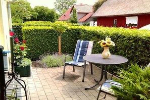 Eine Terrasse mit einem runden Tisch und zwei Stühlen, umgeben von ordentlich geschnittenen Hecken und Topfpflanzen.