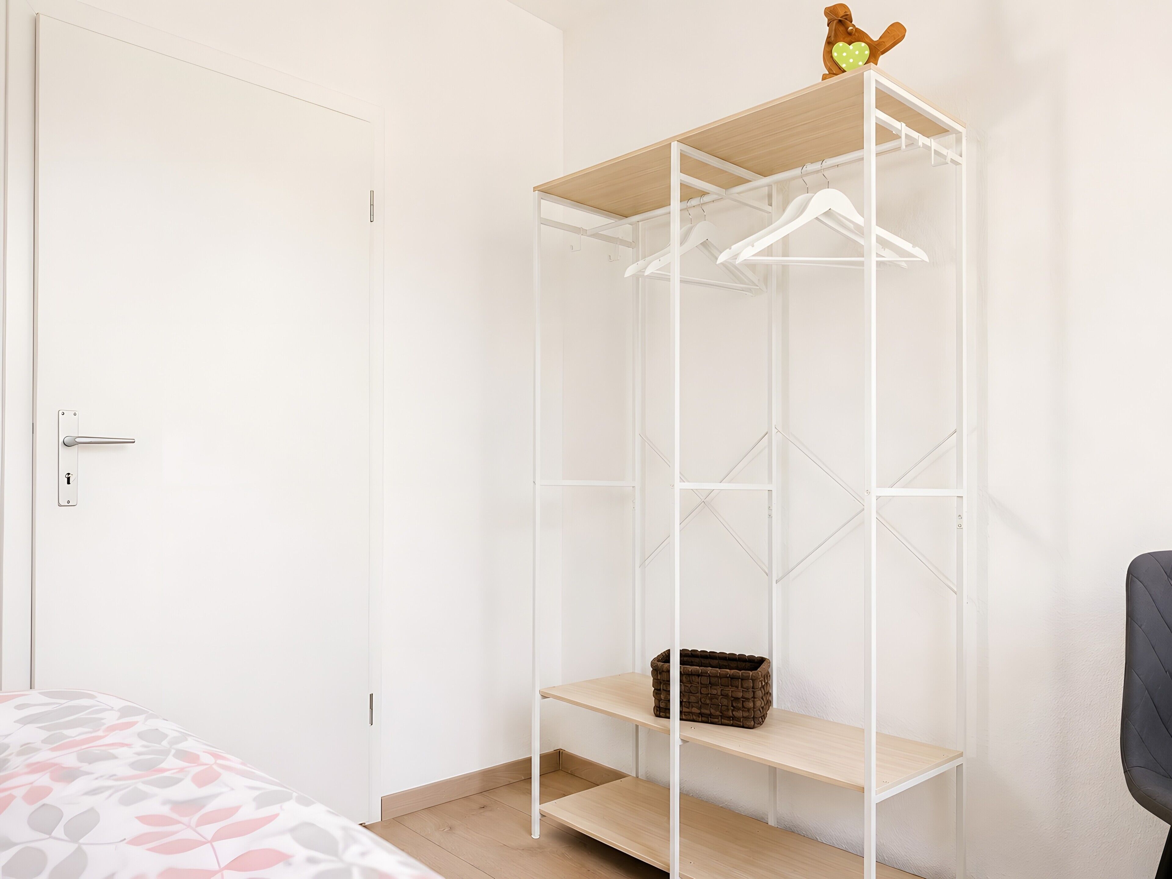 Ein Zimmer mit einer weißen Tür, einem Bett mit gemustertem Überwurf, einem weißen Kleiderschrank mit Regalen und einem kleinen braunen Korb auf einem der Regale.
