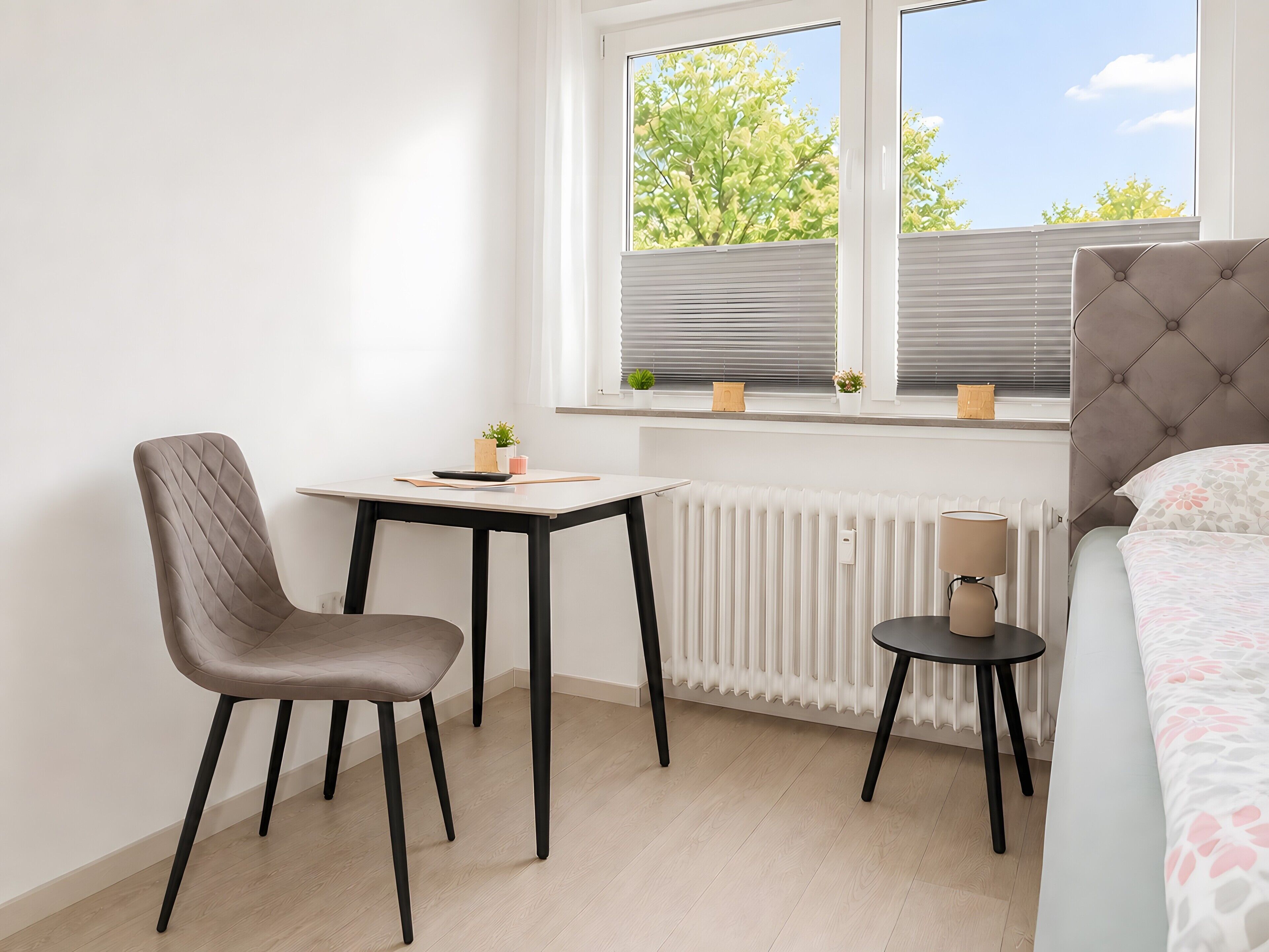 Une pièce comprenant un lit, une chaise, une petite table, un radiateur et une fenêtre avec des stores.