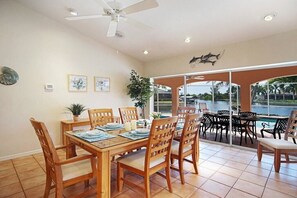 A dining area with a table set for four, a ceiling fan, and a view of a swimming pool through glass doors. - CCVS - Villa Blue Lagoon (Cape Coral)