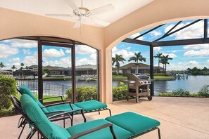 A covered outdoor area with lounge chairs, a grill, and a view of a lake and houses. - CCVS - Villa Blue Lagoon (Cape Coral)