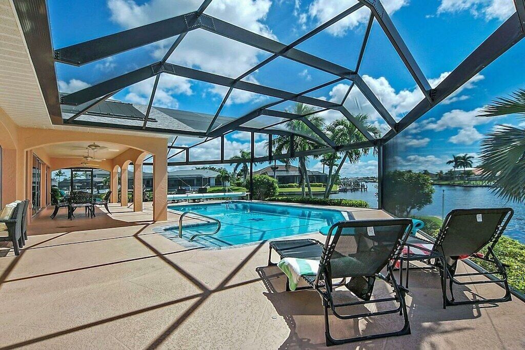 A covered outdoor swimming pool area with lounge chairs, a pool table, and a view of a lake.