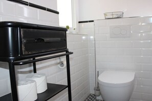 A bathroom with a white toilet, a black shelf with toilet paper, and a shower area. - Holiday apartment with 1 bedroom (Heeslingen)