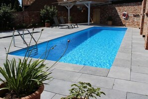 A rectangular swimming pool with clear blue water, surrounded by a paved patio with potted plants and a brick wall in the background. - Holiday apartment with 1 bedroom (Heeslingen)