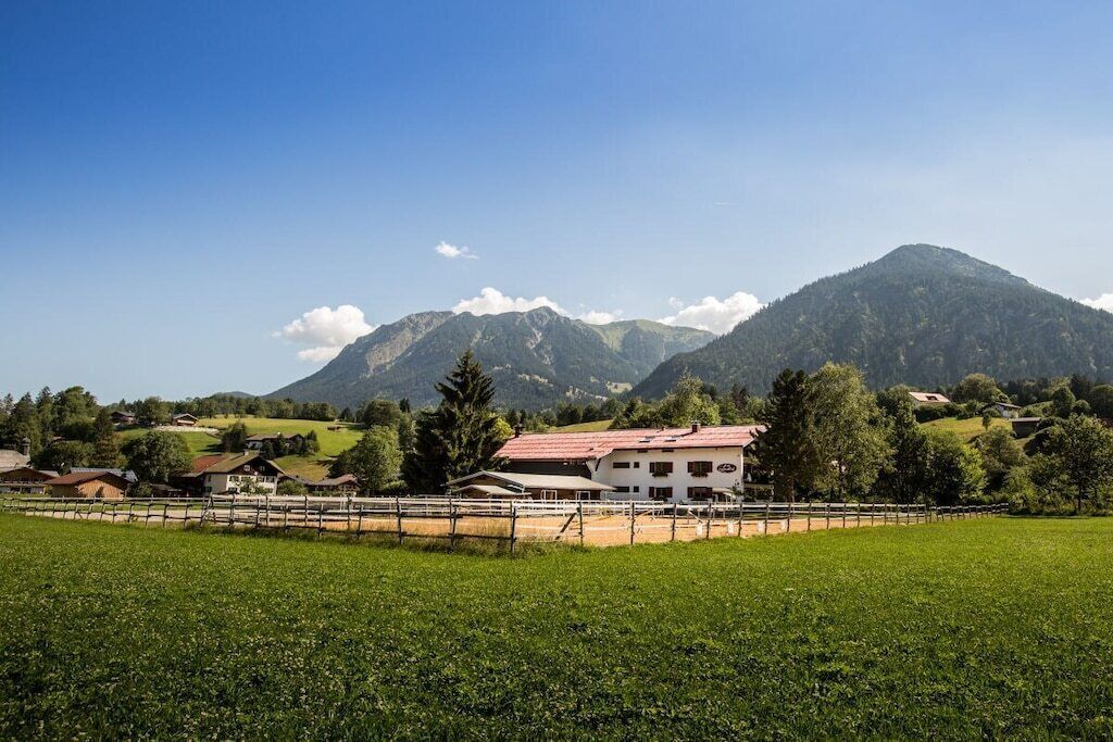 Paisatge rural amb un edifici blanc amb teulada vermella, envoltat de camps verds i muntanyes al fons.