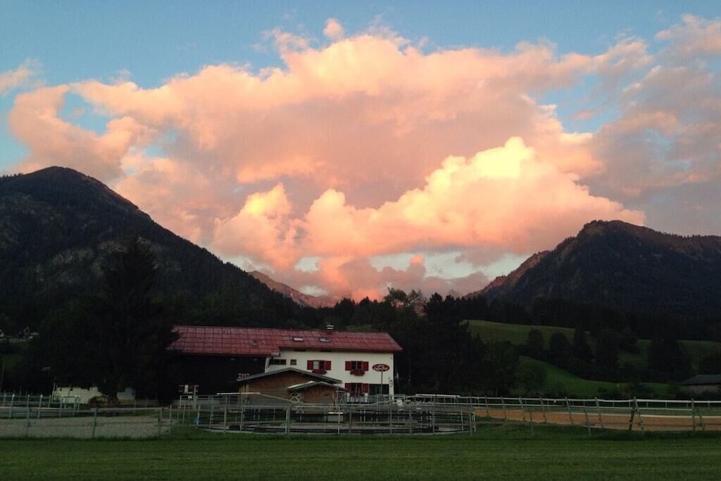 Ein Landhaus mit rotem Dach, umgeben von einem eingezäunten Bereich und Bergen im Hintergrund.