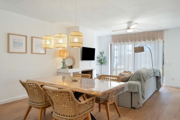 A modern living room with a wooden dining table, wicker chairs, a flat-screen TV, and a ceiling fan.