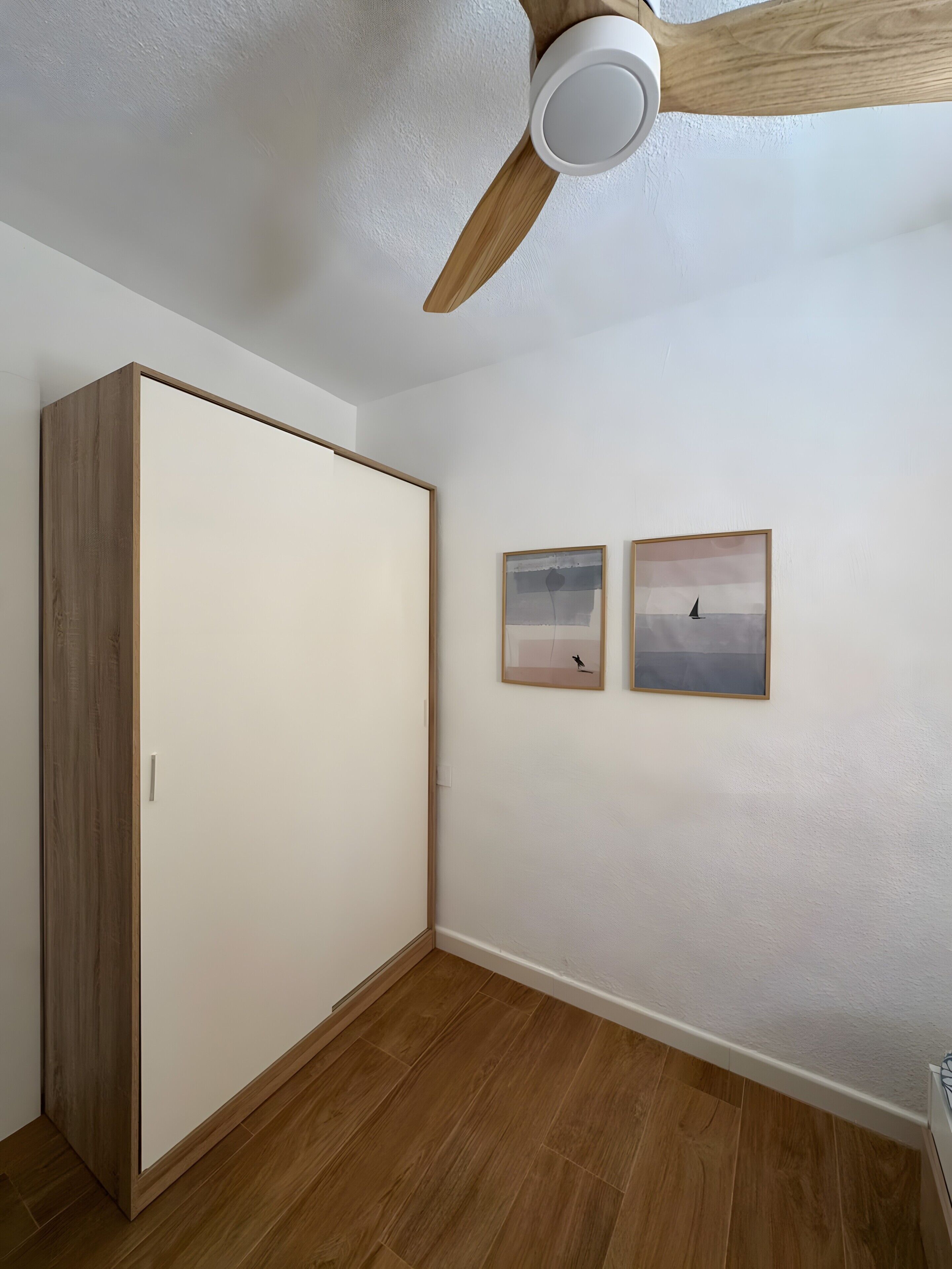 Una habitación con un armario de madera, un ventilador de techo y dos cuadros en la pared.