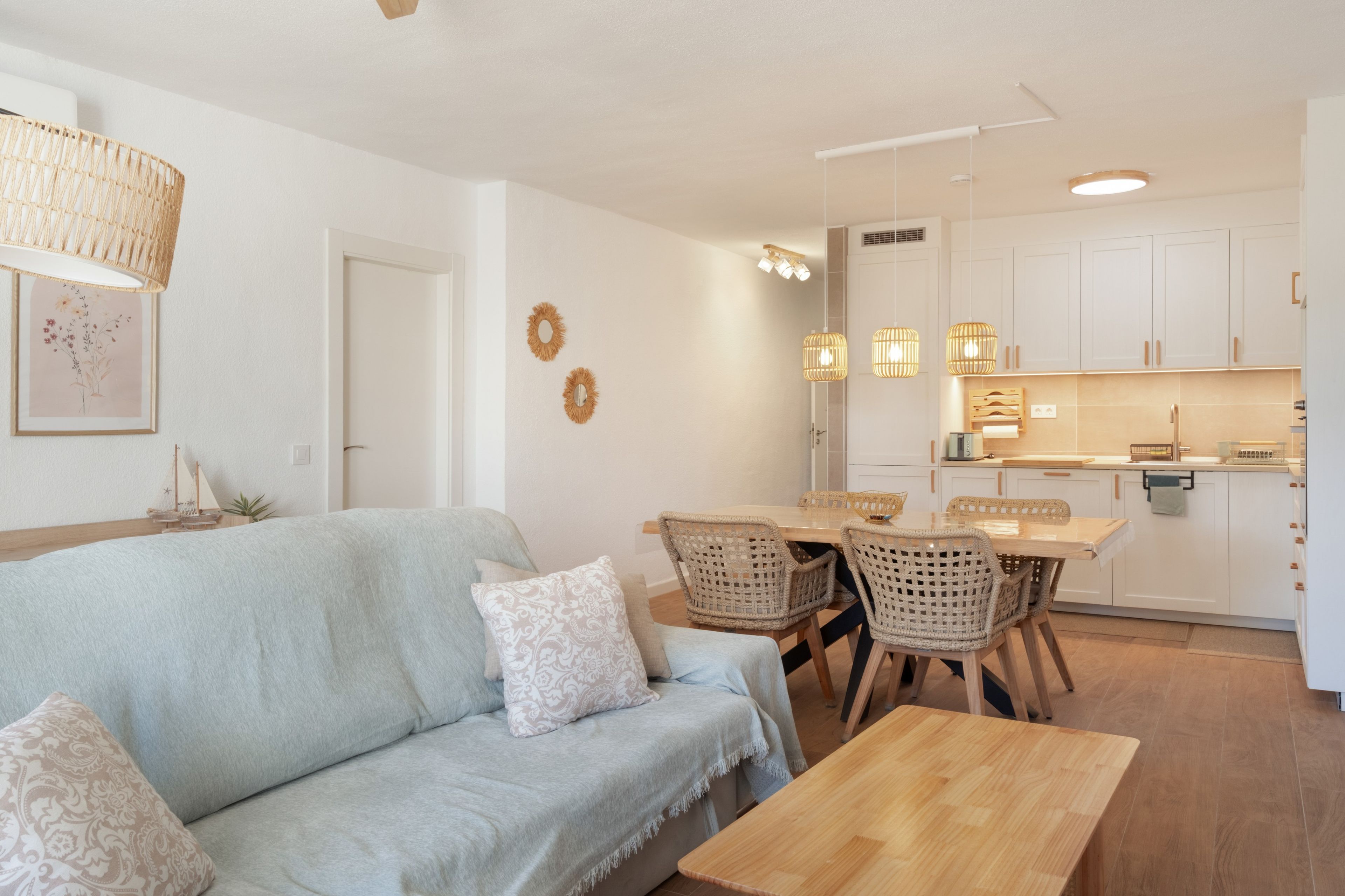 Ein modernes Wohnzimmer mit einem hellblauen Sofa, einem Holztisch und Korbstühlen. Es gibt einen Essbereich mit einem Holztisch und Stühlen, sowie eine Küche mit weißen Schränken und Pendelleuchten.