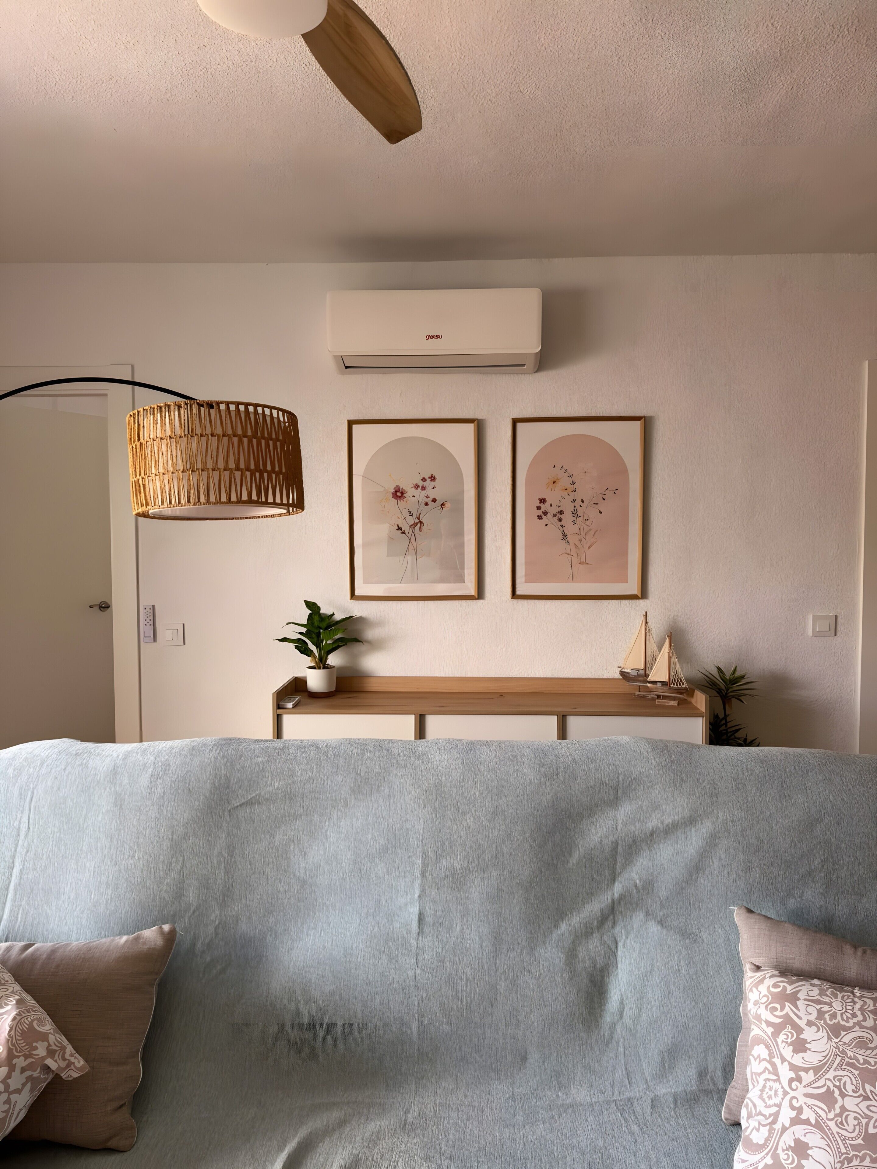 A bedroom with a bed, a ceiling fan, a wall-mounted air conditioner, two framed floral artworks, a wooden bench, and a potted plant.
