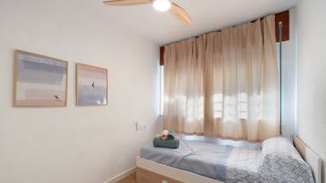 Ein Schlafzimmer mit einem Einzelbett, einem Deckenventilator, zwei Wandbildern und einem Fenster mit Vorhängen.