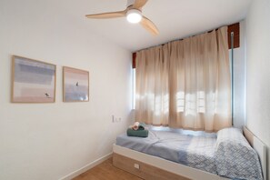 Een slaapkamer met een enkel bed, een plafondventilator, twee aan de muur hangende foto's en een raam met gordijnen.