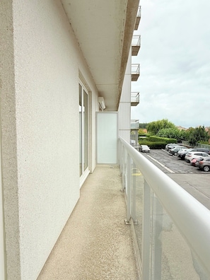 A narrow balcony with a concrete floor and a white railing, overlooking a parking area with several cars and a green landscape in the background. - Lovely Apartment for 6 Guests with Beautiful Water Views Baby Cot Included (Knokke-Heist)