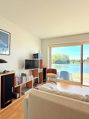 A living room with a sofa, a television, a bookshelf, and a view of a lake through large windows. - Lovely Apartment for 6 Guests with Beautiful Water Views Baby Cot Included (Knokke-Heist)