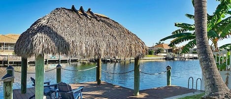 Una estructura con techo de paja, con asientos, una terraza de madera y una masa de agua al fondo.