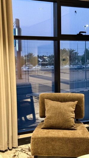 Quarto com uma janela ampla, um sofá com almofada e vista para a rua com palmeiras e prédios.