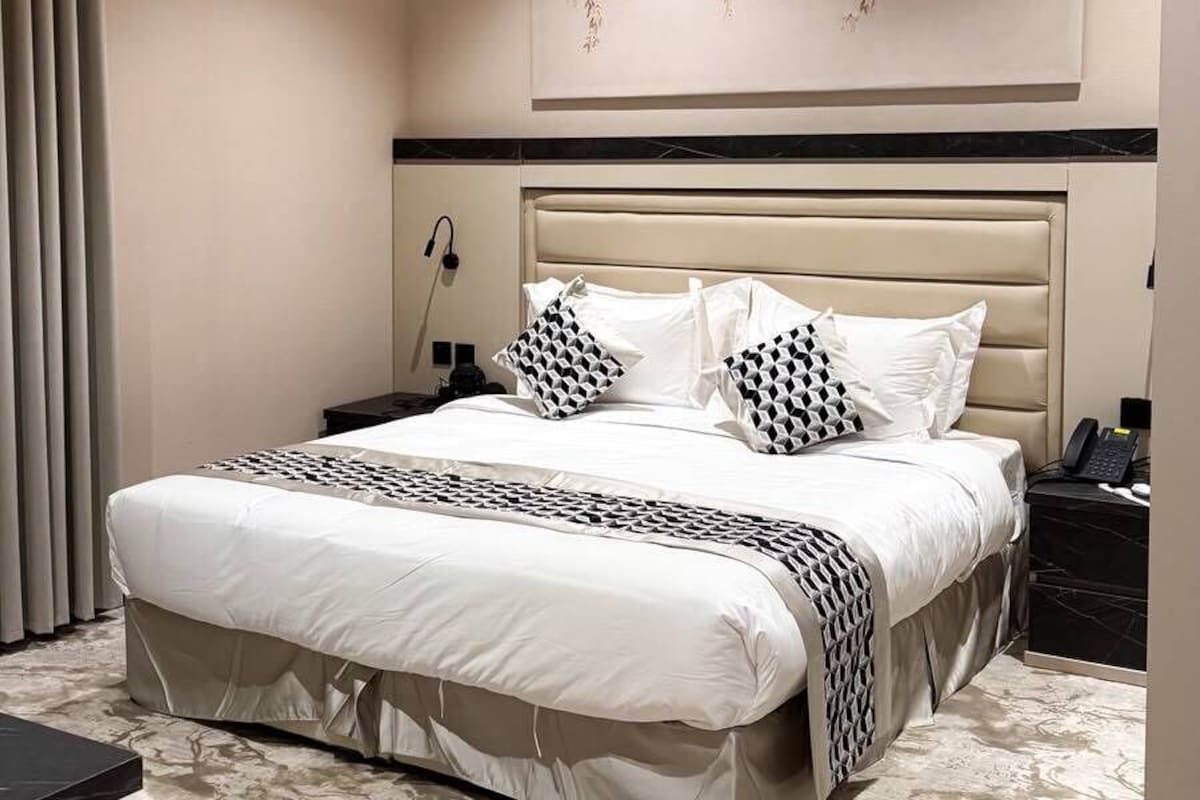 Quarto de hotel com uma cama grande, uma mesa de cabeceira, ar condicionado fixado na parede e um objeto decorativo sobre a cama.