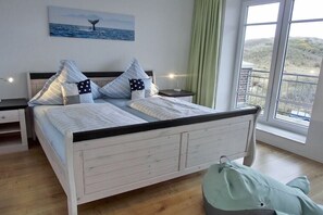 A bedroom with a bed, bedside tables, a nightstand, a window with a view, and a painting on the wall. - Beach Villa Kleckerburg (Langeoog)