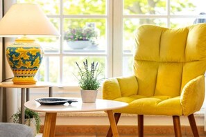 Une pièce avec un fauteuil jaune, une table avec une lampe et une fenêtre donnant sur un paysage verdoyant.