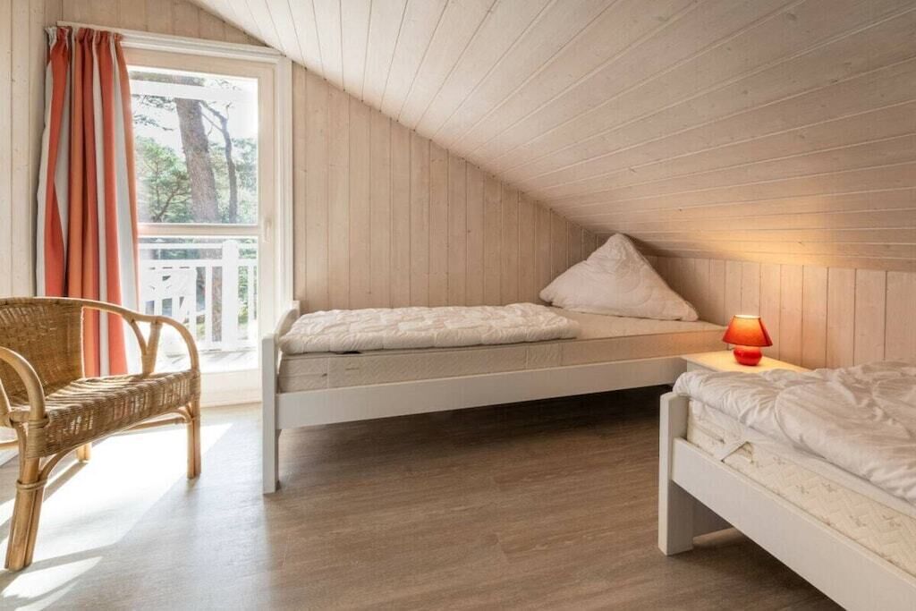 A bedroom with two single beds, a wicker chair, a window with curtains, and a wooden ceiling.