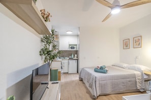 A compact bedroom with a bed, bedside table, and a small desk with a plant. A kitchenette with a microwave and cabinets is visible in the background. - Cozy Studio Apartment in Cala Del Pino with Wi-Fi and Air Conditioning (Cartagena)