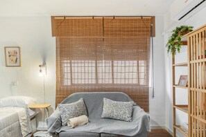 Une pièce avec un canapé gris, une petite table d’appoint, une lampe, une bibliothèque en bois et une grande fenêtre avec des stores en bambou.