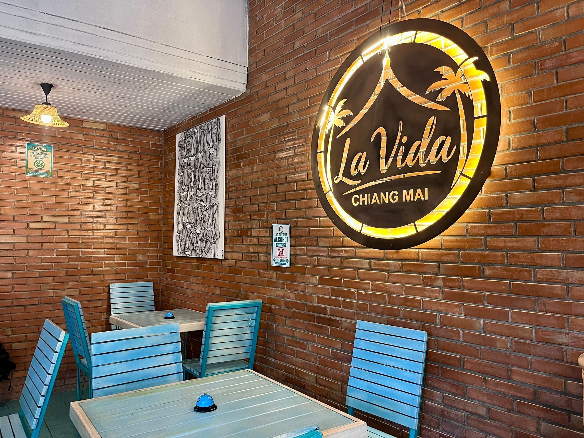 A restaurant interior with a brick wall, a sign reading 'La Vida Chiang Mai', and blue chairs around a table.