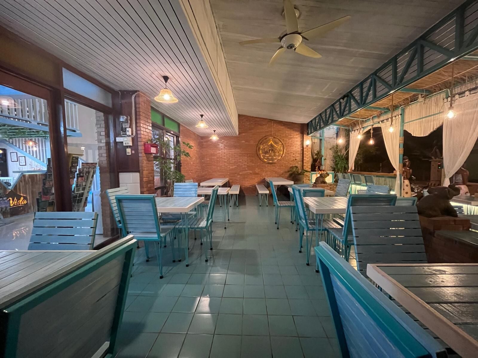 An indoor dining area with tables and chairs, a ceiling fan, and a brick wall with a circular emblem.