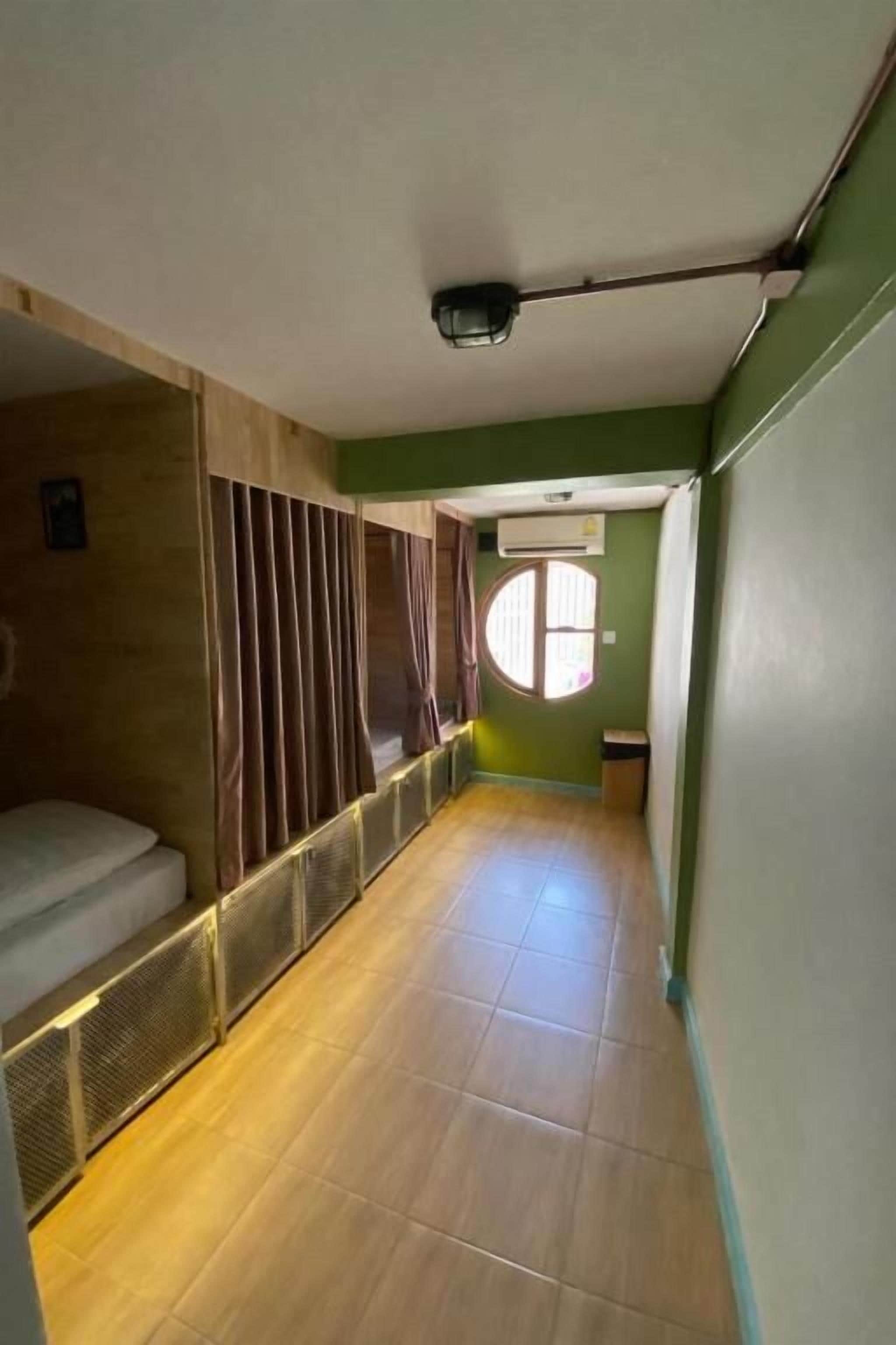 Un estrecho pasillo con una cama a la izquierda, una ventana con cortinas y una puerta al final.