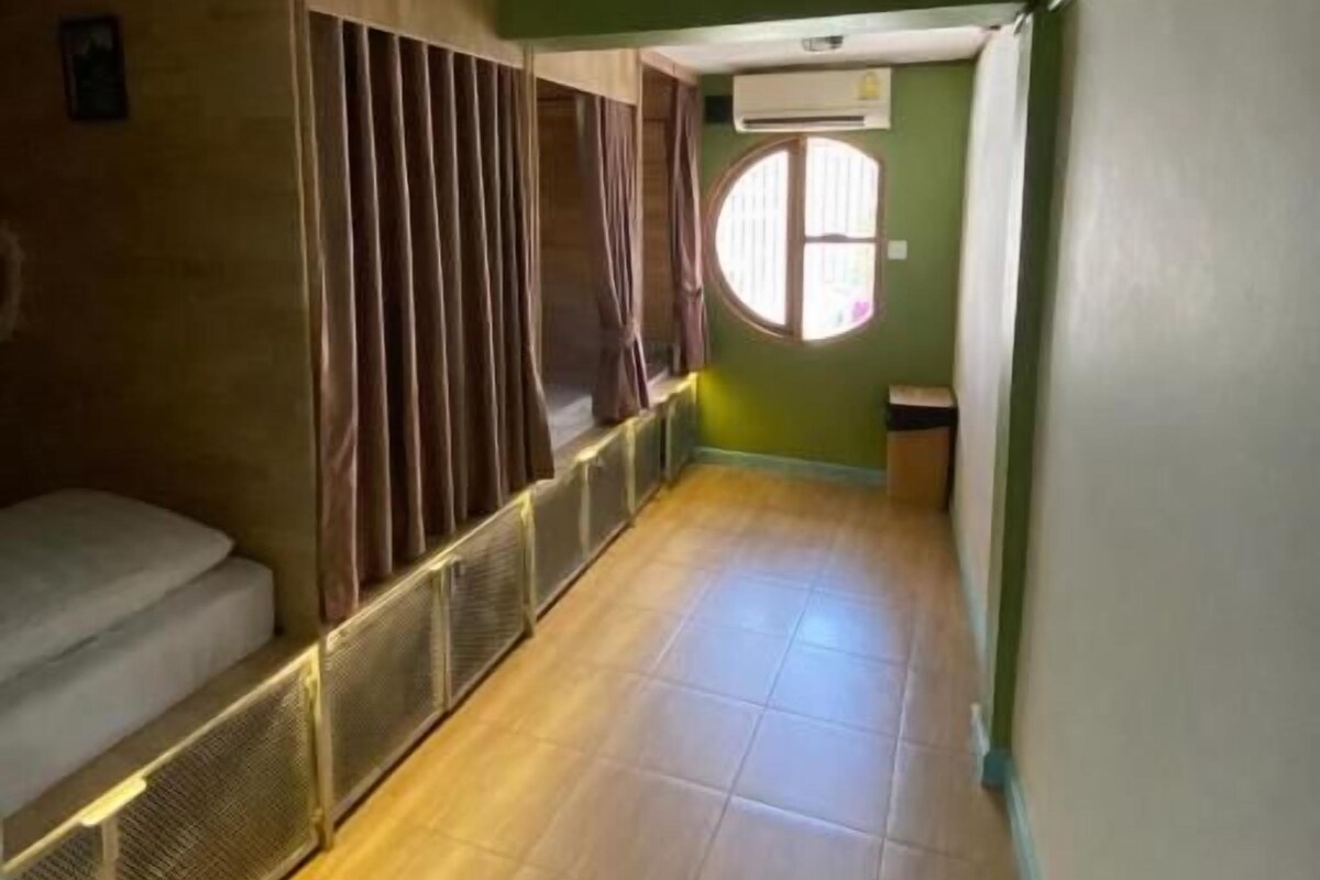 En smal hall med en säng på vänster sida, ett fönster med gardiner och en dörr längst ner.