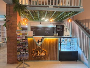 Ett kafé som heter ”La Vida”, med en bardisk i trä, en menytavla och en trappa som leder uppåt till höger.