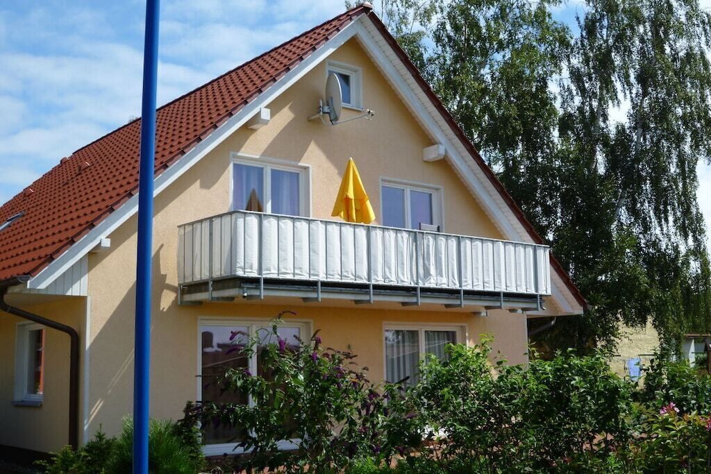 Uma casa de dois andares com telhado de telhas vermelhas, uma varanda com grades brancas e um guarda-sol amarelo.