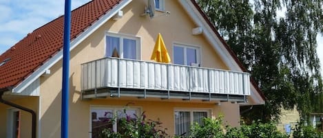 Uma casa de dois andares com telhado de telhas vermelhas, uma varanda com grades brancas e um guarda-sol amarelo.