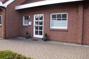 A red brick building with a white door and windows, a paved entrance, and two potted plants. - Pennekamp Carolinensiel (Carolinensiel)
