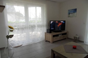 A living room with a television on a wooden stand, a glass vase with a flower, and a table with a red object on it. - Holiday apartment with 2 bedrooms (Zinnowitz)