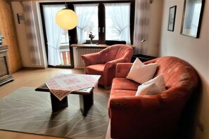 A living room with a sofa, armchair, and a coffee table. There is a window with curtains, a radiator, and a hanging light fixture. - Holiday apartment with 1 bedroom (Titisee- Neustadt)