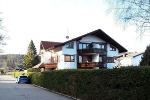 A two-story residential building with a balcony, a car parked in front, and a hedge along the driveway. - Holiday apartment with 1 bedroom (Titisee- Neustadt)