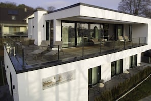 A modern two-story building with a flat roof, large windows, and a balcony with outdoor seating.