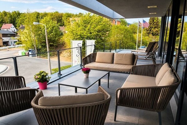 An outdoor seating area with wicker furniture and a glass table, overlooking a street with houses and greenery.